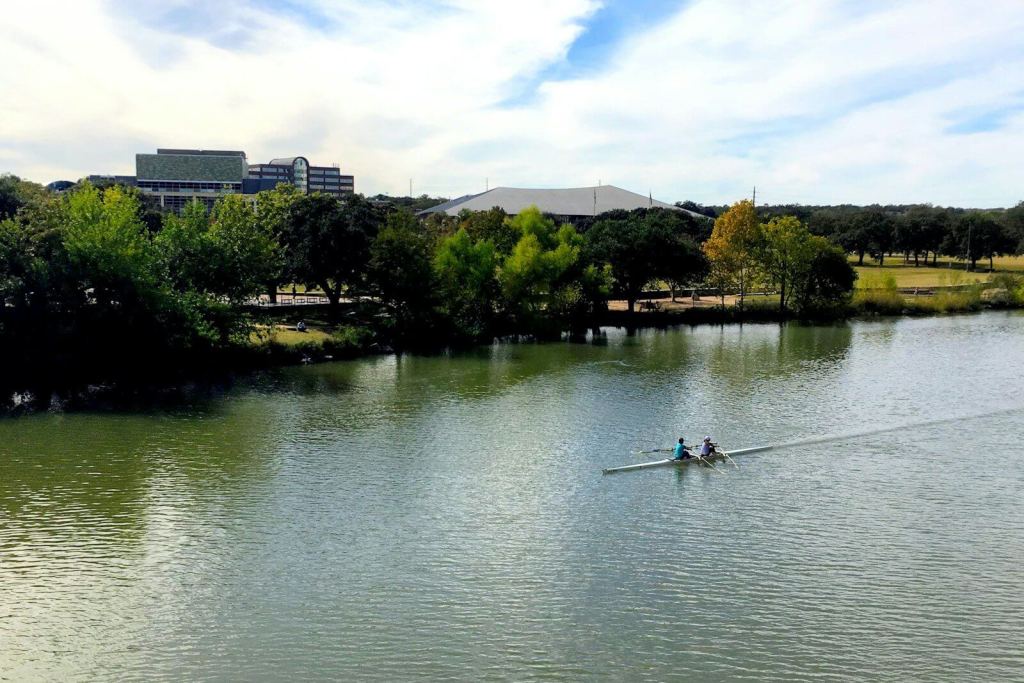 Two people rowing a scull on a river past a tree-lined park.