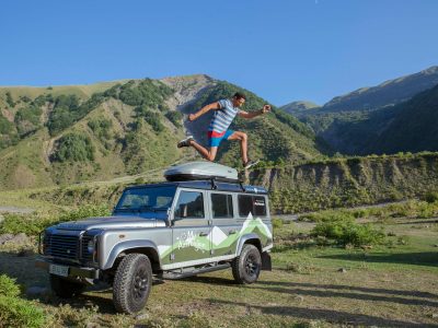 Man jumps on SUV roof in a beautiful mountainous landscape, symbolizing adventure and thrill.